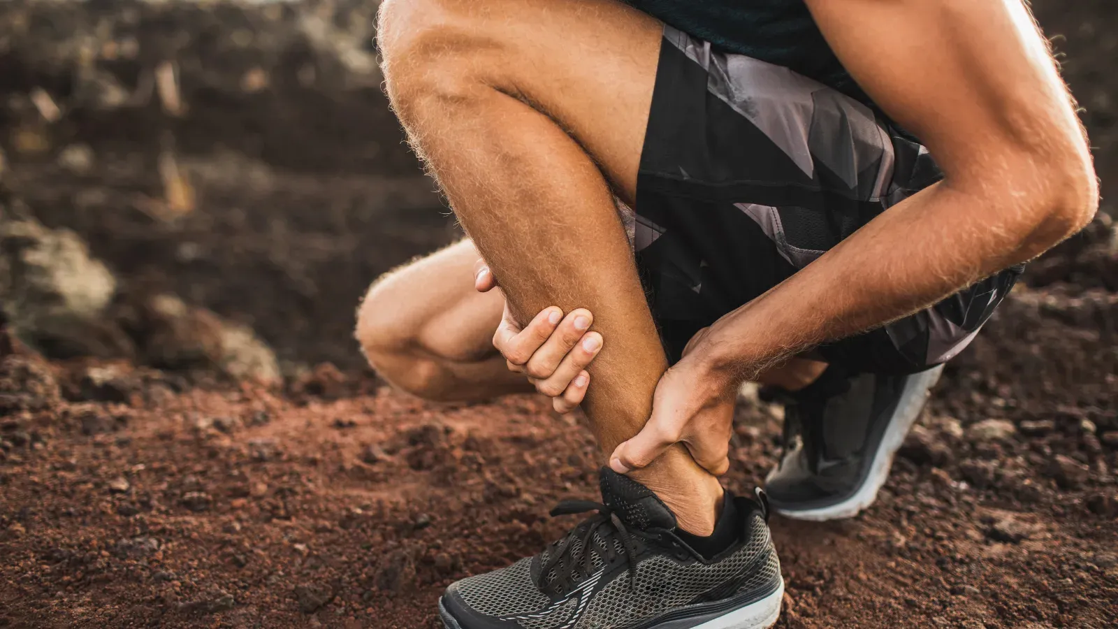 A runner crouching on a trail, gripping the back of their ankle