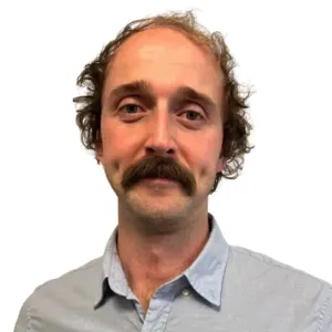 Head and shoulders portrait of a physiotherapist with curly hair and a moustache, in a light blue shirt