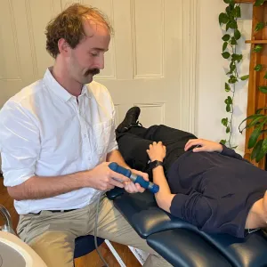 A physiotherapist holding a shockwave handpiece to the forearm of a patient lying on a treatment table