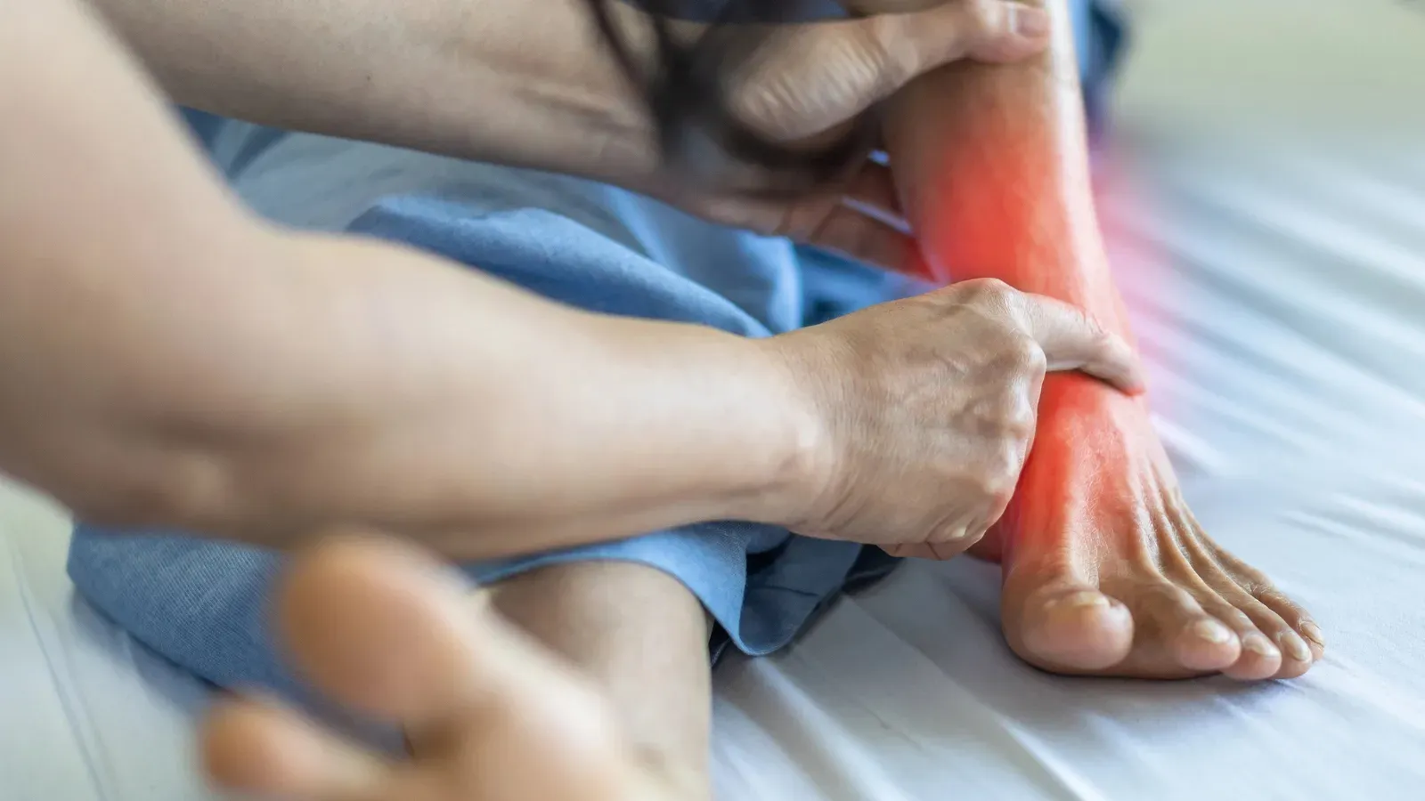 A person sitting on a bed holding their ankle, the joint highlighted in red