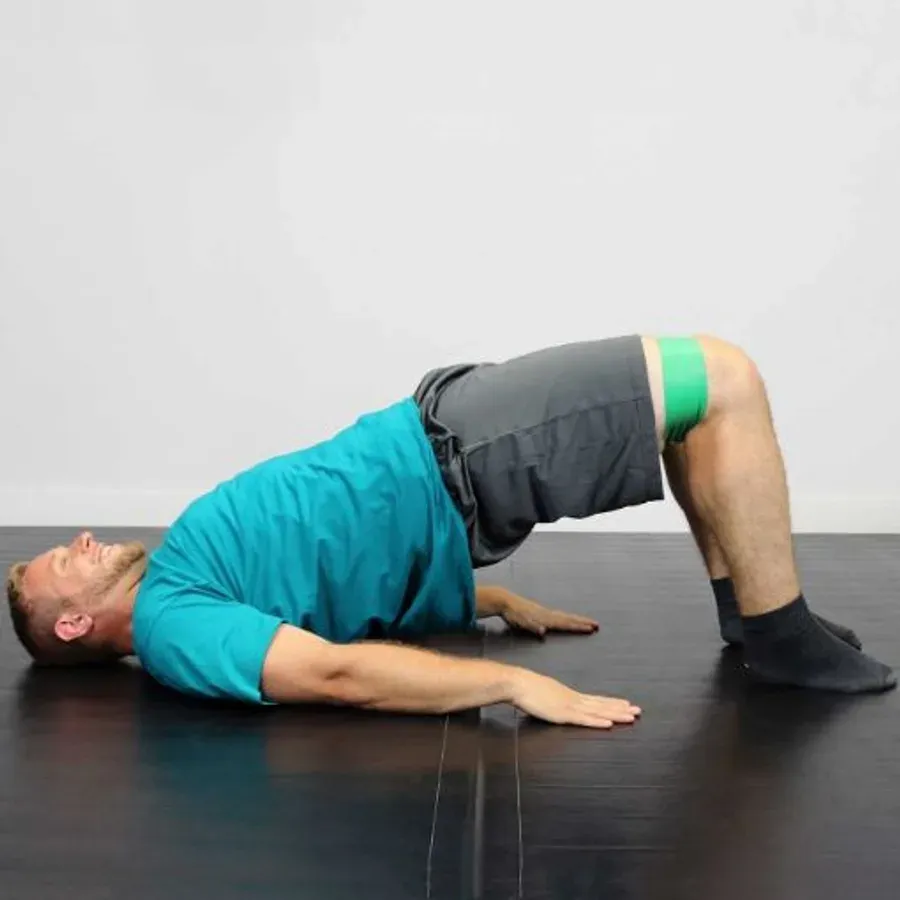A man in a glute bridge on the floor with a green band loop around his knees