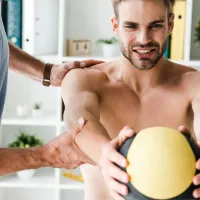 A physiotherapist steadying a man's shoulder and elbow as he holds a medicine ball out in front of him