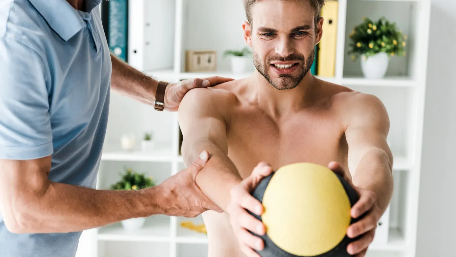 A man holding a weighted ball while a therapist guides his shoulder