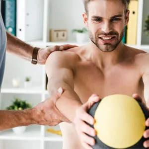 A physiotherapist steadying a man's shoulder and elbow as he holds a medicine ball out in front of him