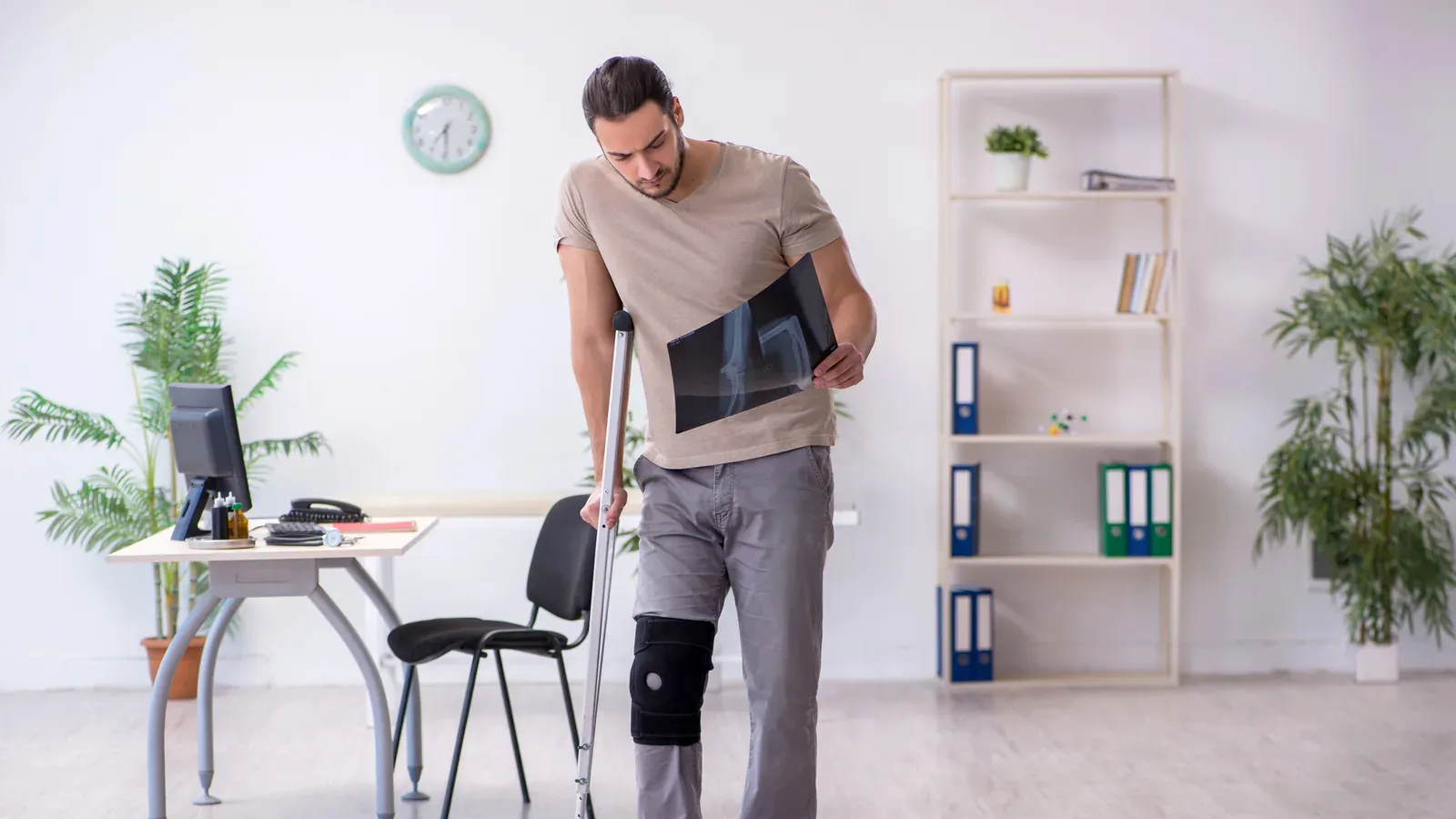 A man on a crutch wearing a knee brace, studying an X-ray in a consulting room