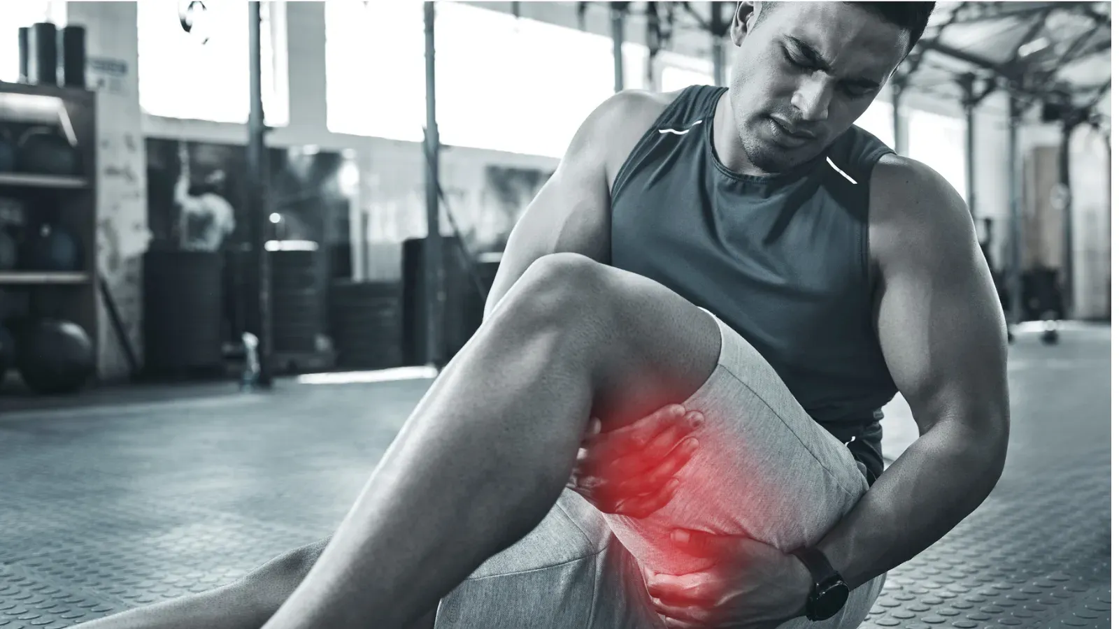 A man sitting on a gym floor gripping the back of his thigh, the hamstring highlighted in red