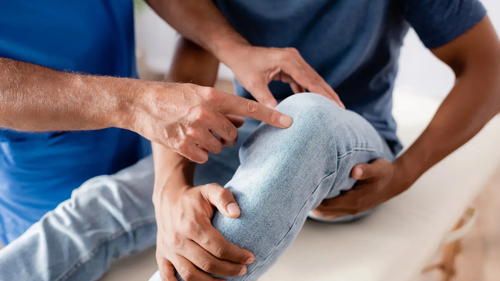 A physiotherapist examining a seated patient's knee, pointing to the joint line