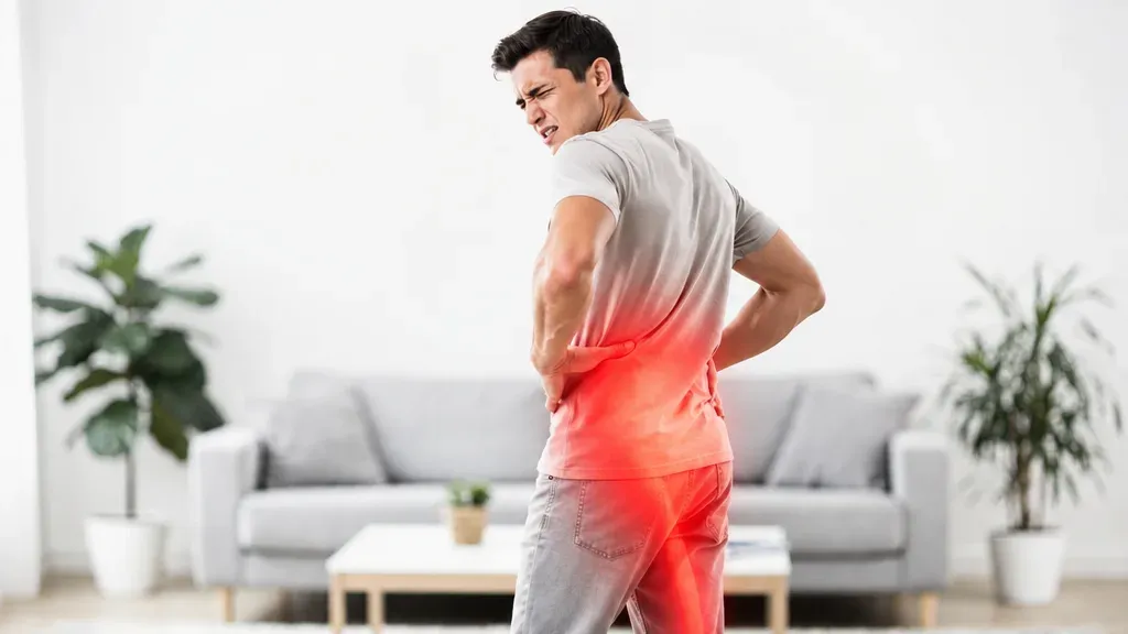 A man holding his lower back and wincing, pain highlighted in red across the base of the spine
