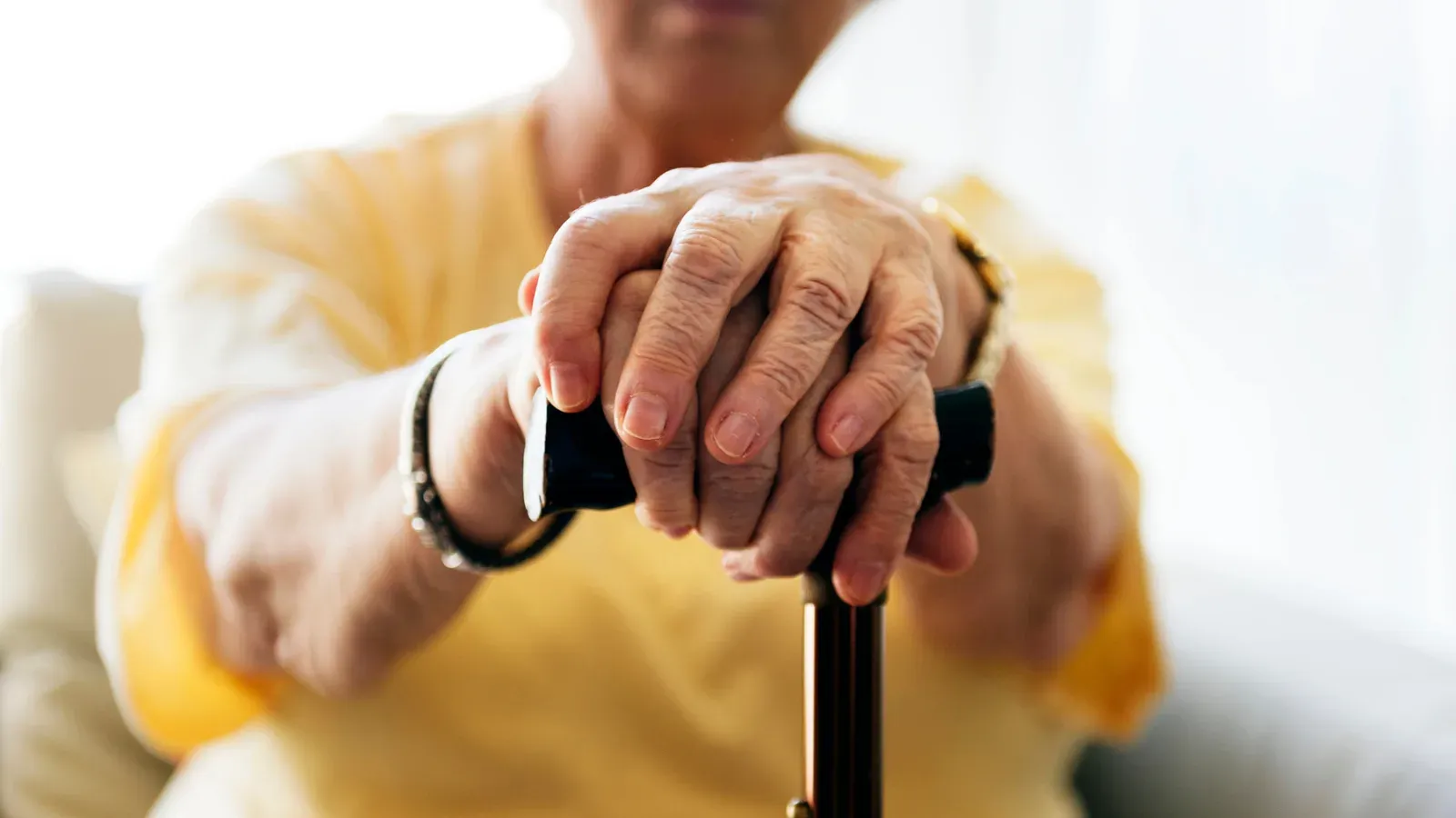 An older person's hands resting one over the other on the handle of a walking stick