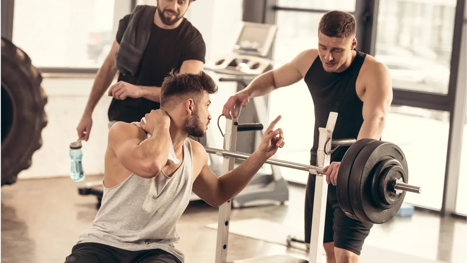 A man on a gym bench reaching for his shoulder while two training partners look on