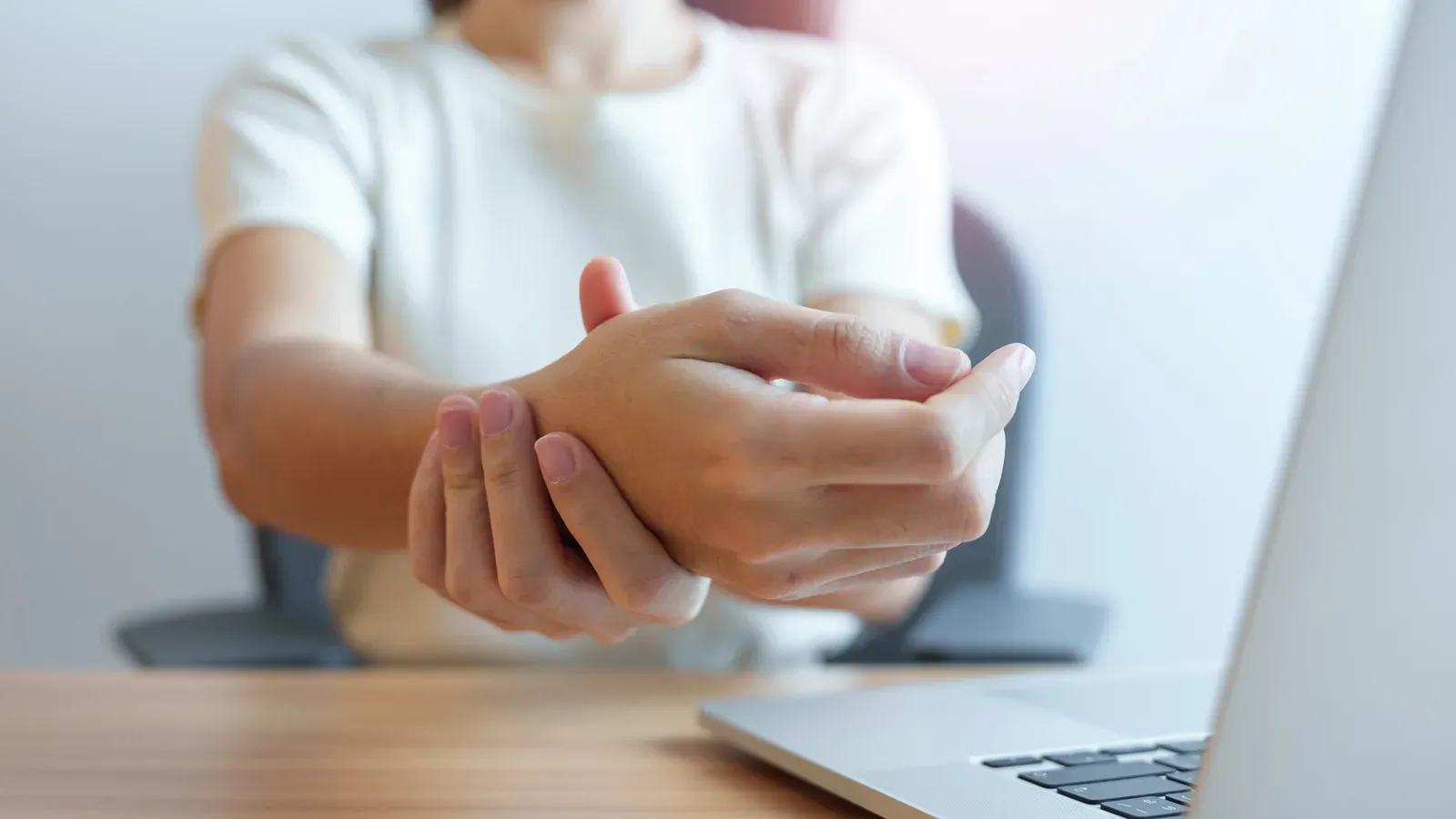 A person at a desk holding their wrist, a laptop open in front of them
